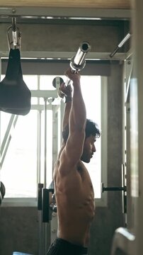 Athletic man performing an overhead barbell press in a gym, side profile by a window, focused strength training.