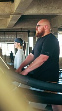 Two people exercising on treadmills in an industrial-style gym, focused on cardio training near large windows.