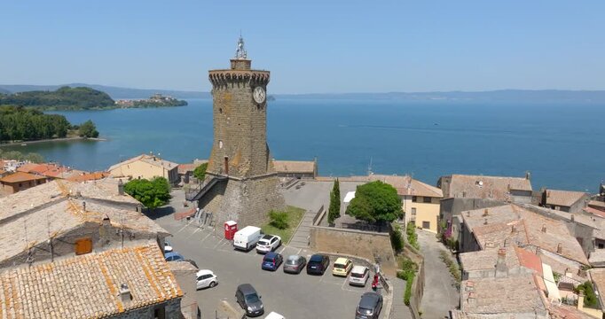Aerial view of the clock tower, a car park and the old stone houses of Marta overlooking Lake Bolsena, with a wooded shore on the horizon, in the province of Viterbo, in Lazio, Italy, on a sunny day