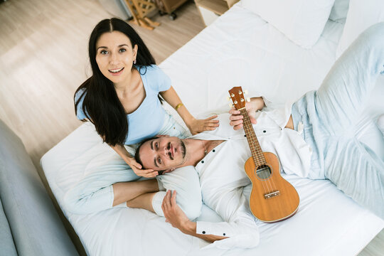 Smiling couple relaxing on a bed at home; he rests with a ukulele while she kneels beside him, both looking up at the camera in a cozy, bright bedroom