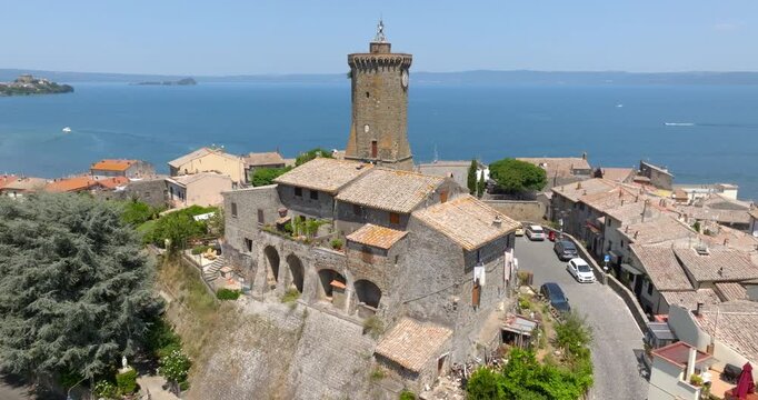 Aerial view of the clock tower and an old stone building with arches in the historic center of Marta, with a road, Lake Bolsena and Bisentina island, in the province of Viterbo, in Lazio, Italy.