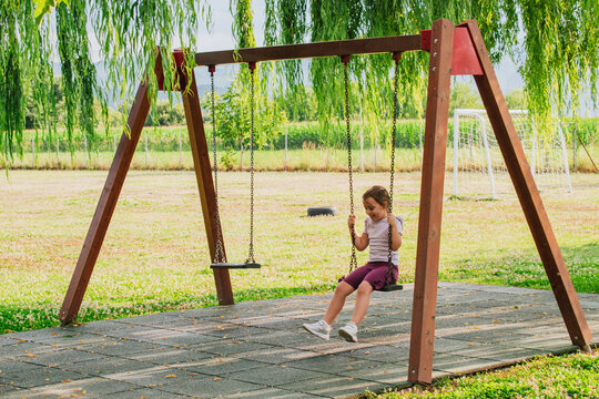 Happy little girl swinging on wooden chain swing set at outdoor park playground under green trees on sunny day for childhood active vacation stock photo