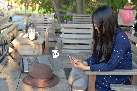 Woman smiling while using smartphone at outdoor wooden table, with laptop, coffee, and hat, enjoying mobile phone communication and relaxed lifestyle in garden.