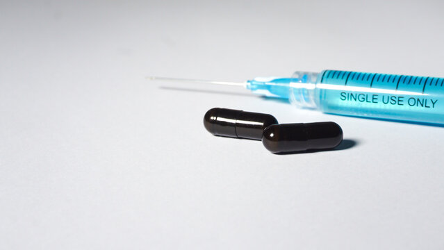 Disposable medical syringe contain blue liquid and two black capsules on white surface. 