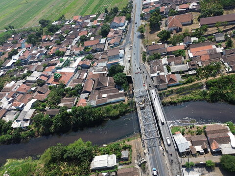 Aerial view of a steel truss bridge spanning a river in a residential area with tiled rooftops, green fields, and vehicles on the road, Probolinggo Regency, East Java, Indonesia.