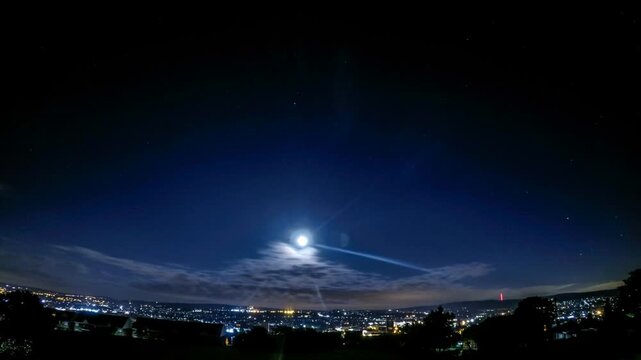Nighttime time-lapse footage featuring a bright moon shining in a deep blue sky as clouds swiftly pass overhead above the distant streetlights and urban landscape of Blackburn, Lancashire, England.
