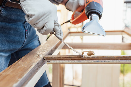Adult carpenter wearing protective leather gloves, using a heat gun and a wood scraper to remove paint from an old wooden window. Construction industry, carpentry. Restoration. 