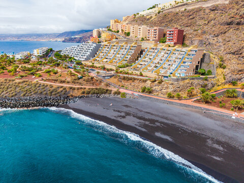 Aerial view of Tenerife coastline featuring modern terraced apartment buildings overlooking a volcanic black sand beach with turquoise waters in Santa Cruz de Tenerife, Canary Islands, Spain.