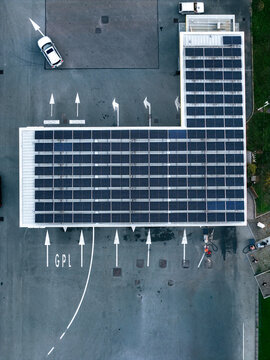 Aerial view of a petrol station featuring a large roof covered in solar panels, with painted road markings and a white car parked on the asphalt in Orbassano, Piedmont, Italy.
