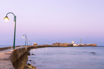 Stone causeway stretching toward San Sebastian Castle lit by street lamps at pre-dawn twilight, surrounded by the ocean in Cadiz, Andalusia, Spain. © nzatravel
