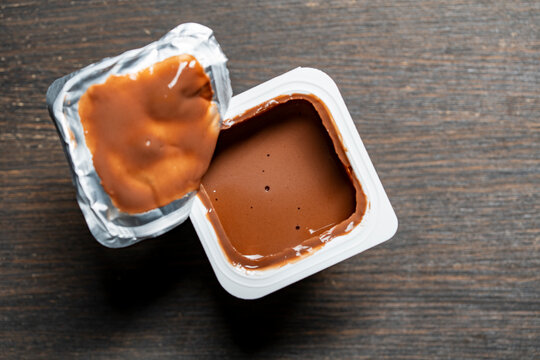 Opened chocolate pudding plastic cup on wooden background. Top view of open white plastic container with sweet chocolate mousse or yogurt. Creamy dairy dessert with opened foil lid on dark table