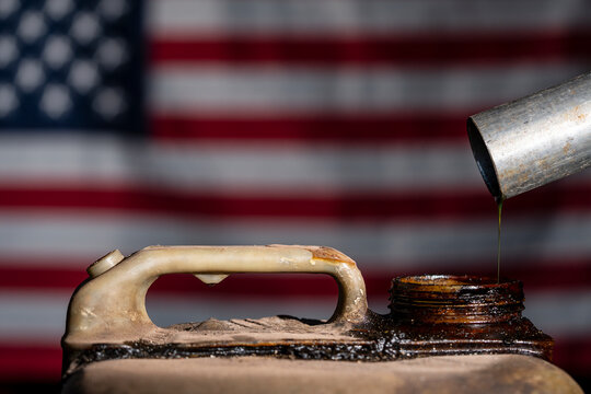 Pouring oil into old rusty canister with American flag. Fuel pouring from metal pipe into dirty jerrycan. Defocused USA flag background for american oil industry energy crisis or fuel policy concept