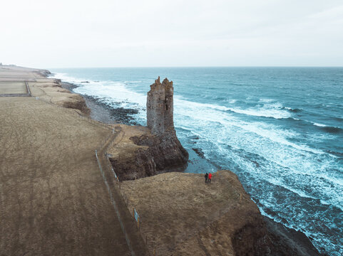 Aerial view of Old Keiss Castle standing on a rugged cliff edge overlooking the sea with two people standing nearby on the grassy coast in Keiss, United Kingdom.