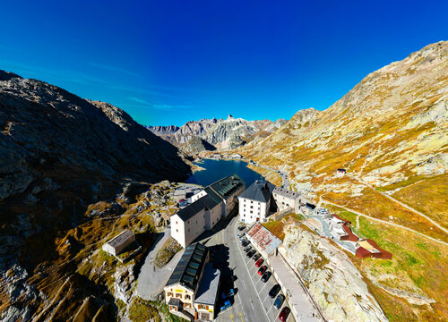Aerial view of Great St Bernard Hospice, alpine lake, winding road, and parked cars in the rugged mountains of Bourg-Saint-Pierre, Valais, Switzerland.