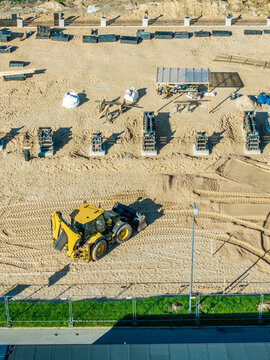 Aerial view of a yellow backhoe loader working on a sandy construction site with concrete foundation forms and a temporary shelter in Ragana, Sigulda Municipality, Latvia.