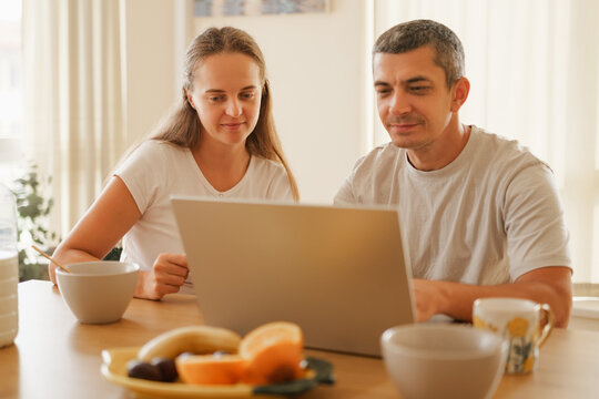 Focused couple sitting at a kitchen table during breakfast while looking together at a laptop screen.