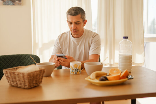Focused man holding and using a smartphone while sitting alone at a dining table during breakfast in a bright kitchen.