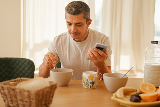 Focused man holding a smartphone in one hand while eating breakfast with a spoon at a dining table in a bright kitchen.