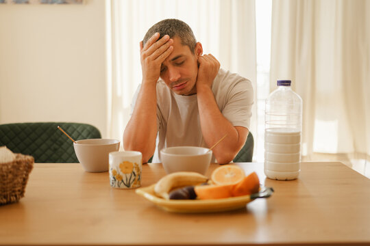 Tired man holding his head in hand while sitting alone at a breakfast table feeling unwell or stressed in a bright kitchen.