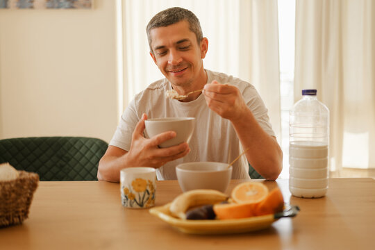 Happy smiling man eating breakfast with a spoon from a bowl sitting at a dining table in a bright kitchen.