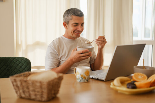 Smiling man eating breakfast from a bowl with a spoon while looking at a laptop screen on a dining table in a bright kitchen.