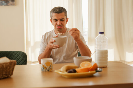 Man with a disgusted grimace holding a bowl and spoon while eating tasteless breakfast at a kitchen table.