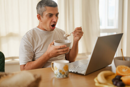 Shocked man with mouth open looking at laptop screen while holding a bowl and spoon during breakfast in kitchen.