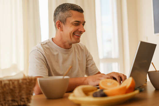 Smiling adult man winking while typing on a laptop computer at a dining table during breakfast in a bright kitchen.