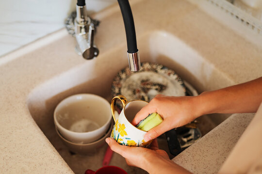Close-up of human hands washing a ceramic cup with a sponge in a kitchen sink filled with dirty dishes.