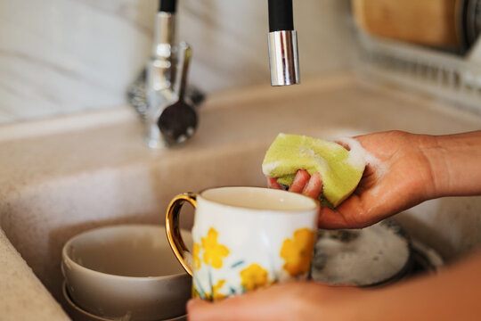 Close-up of human hands holding a foamy sponge and a ceramic mug over a kitchen sink while washing dishes.