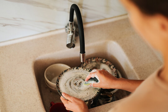 High-angle shot of a person washing a soapy plate with a sponge over a kitchen sink full of dirty dishes.