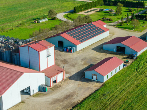 Aerial view of a farm complex with buildings featuring red roofs, solar panels, grain silos, and a tractor on a gravel yard surrounded by green fields in Lode, Sigulda Municipality, Latvia.