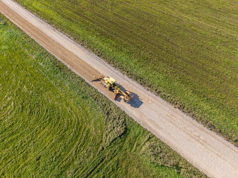 Aerial view of a yellow motor grader leveling and reshaping the surface of a rural gravel road surrounded by green fields in Lode, Sigulda Municipality, Latvia.