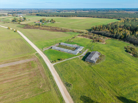 Aerial view of rural farm buildings surrounded by green fields, meadows, and a dirt road in Lode, Sigulda Municipality, Latvia.