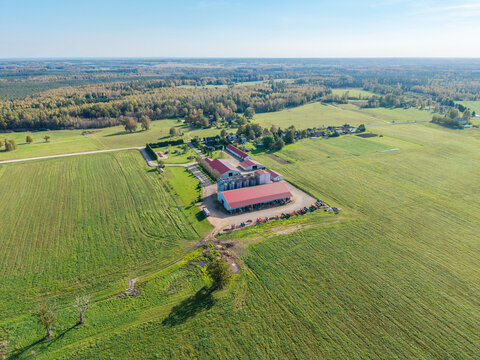Aerial view of a farm complex with red-roofed barns and grain silos surrounded by green fields and forests in Lode, Sigulda Municipality, Latvia.