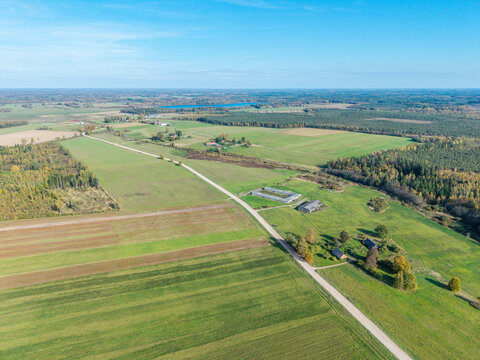 Aerial view of rural landscape in Lode, featuring green agricultural fields, a gravel road, scattered farm buildings, and surrounding forests in Lode, Sigulda Municipality, Latvia.