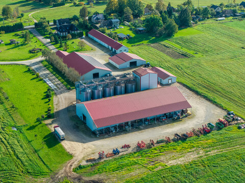 Aerial view of an agricultural farm in Lode featuring grain silos, red-roofed buildings, and machinery parked in a row, surrounded by green fields in Lode, Sigulda, Latvia.