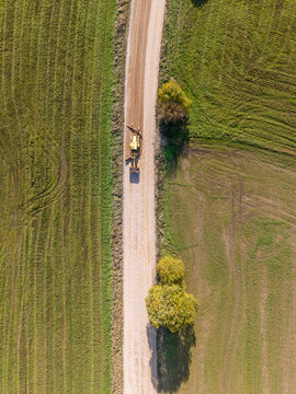 Aerial view of a yellow motor grader leveling and reshaping the surface of a rural gravel road surrounded by green agricultural fields in Lode, Sigulda Municipality, Latvia.