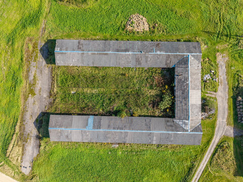 Aerial view of a U-shaped building with a grey tiled roof surrounded by green fields and a dirt path in Lode, Sigulda Municipality, Latvia.