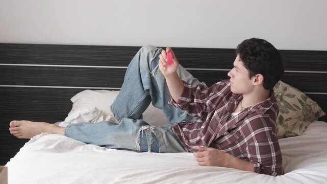 Teenager lounging on bed with gamepad, teen boy in jeans playing video games and raising controller. Lazy weekend and gaming concept, relaxed gamer at home