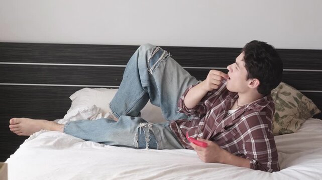 Upset teen gamer losing game, teenager lying on bed with red game controller and frustrated gesture. Video game defeat concept, expressive young player at home