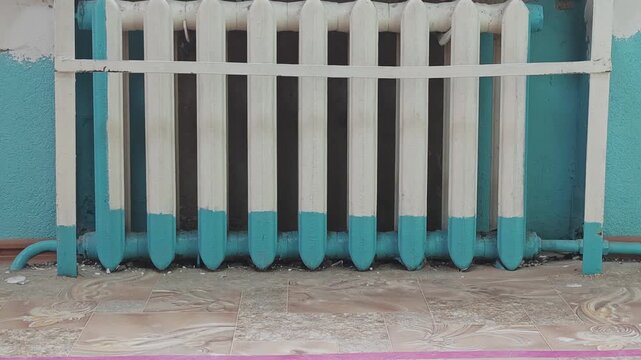 Old cast iron heating radiator against a peeling turquoise wall. Worn tiled floor and decaying interior of an abandoned public building