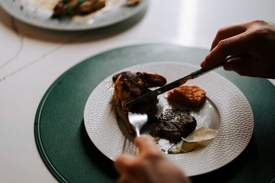 Person cutting a piece of roasted chicken with a knife and fork on a white plate during dinner time