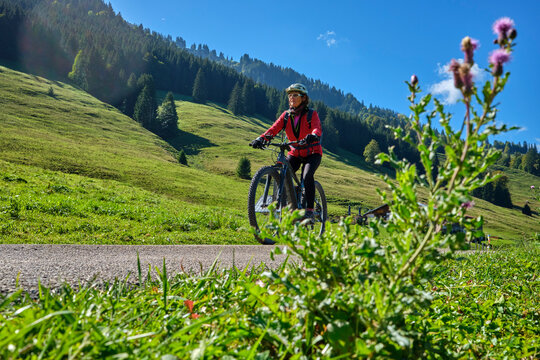 Active senior woman cycling on an electric mountain bike across an alpine meadow with forested slopes and mountain peaks under a blue sky . Allgaeu Alps, Steibis-Oberstaufen, Bavaria, Germany