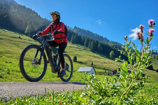 Active senior woman cycling on an electric mountain bike across an alpine meadow with forested slopes and mountain peaks under a blue sky . Allgaeu Alps, Steibis-Oberstaufen, Bavaria, Germany