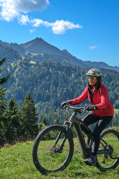 Active senior woman cycling on an electric mountain bike across an alpine meadow with forested slopes and mountain peaks under a blue sky . Allgaeu Alps, Steibis-Oberstaufen, Bavaria, Germany