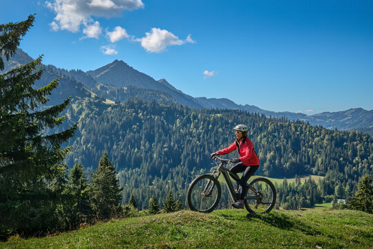 Active senior woman cycling on an electric mountain bike across an alpine meadow with forested slopes and mountain peaks under a blue sky . Allgaeu Alps, Steibis-Oberstaufen, Bavaria, Germany