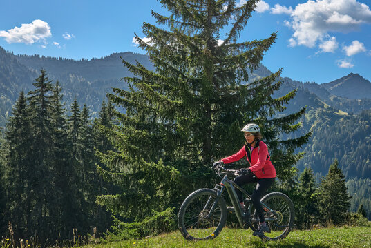 Active senior woman cycling on an electric mountain bike across an alpine meadow with forested slopes and mountain peaks under a blue sky . Allgaeu Alps, Steibis-Oberstaufen, Bavaria, Germany