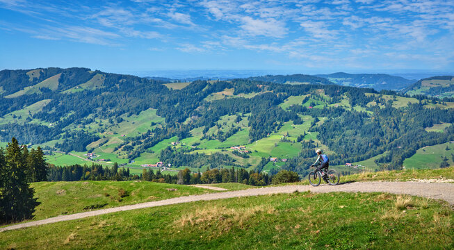 Female mountain biker overlooking a scenic alpine valley with green hills, dense forests and distant villages on a sunny day. Thalkirchdorf, Bavaria, Germany