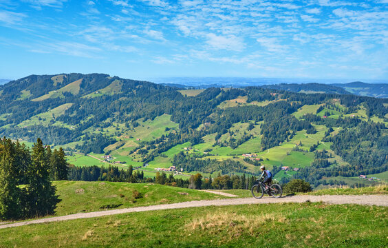 Female mountain biker overlooking a scenic alpine valley with green hills, dense forests and distant villages on a sunny day. Thalkirchdorf, Bavaria, Germany